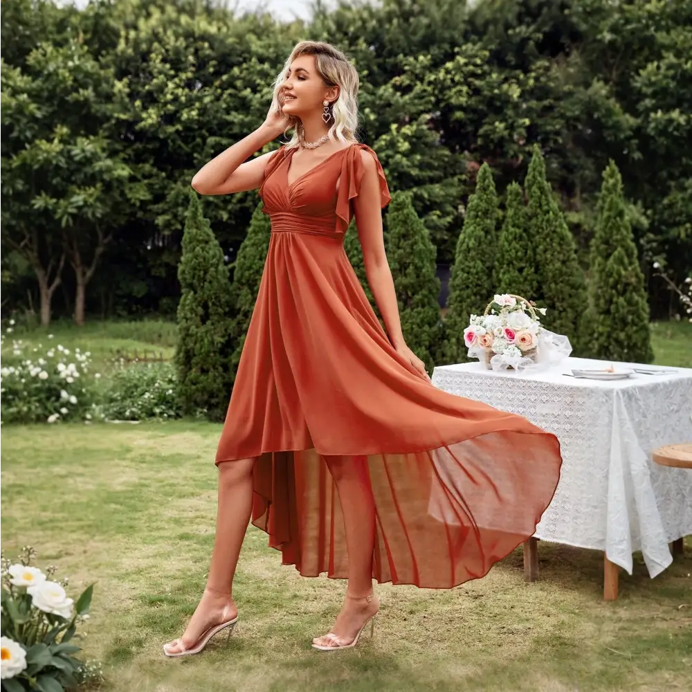 Vue de côté d'une robe cocktail chic originale en terracotta, avec un décolleté en V et des manches courtes, portée par une femme blonde dans un jardin, accompagnée de talons transparents.