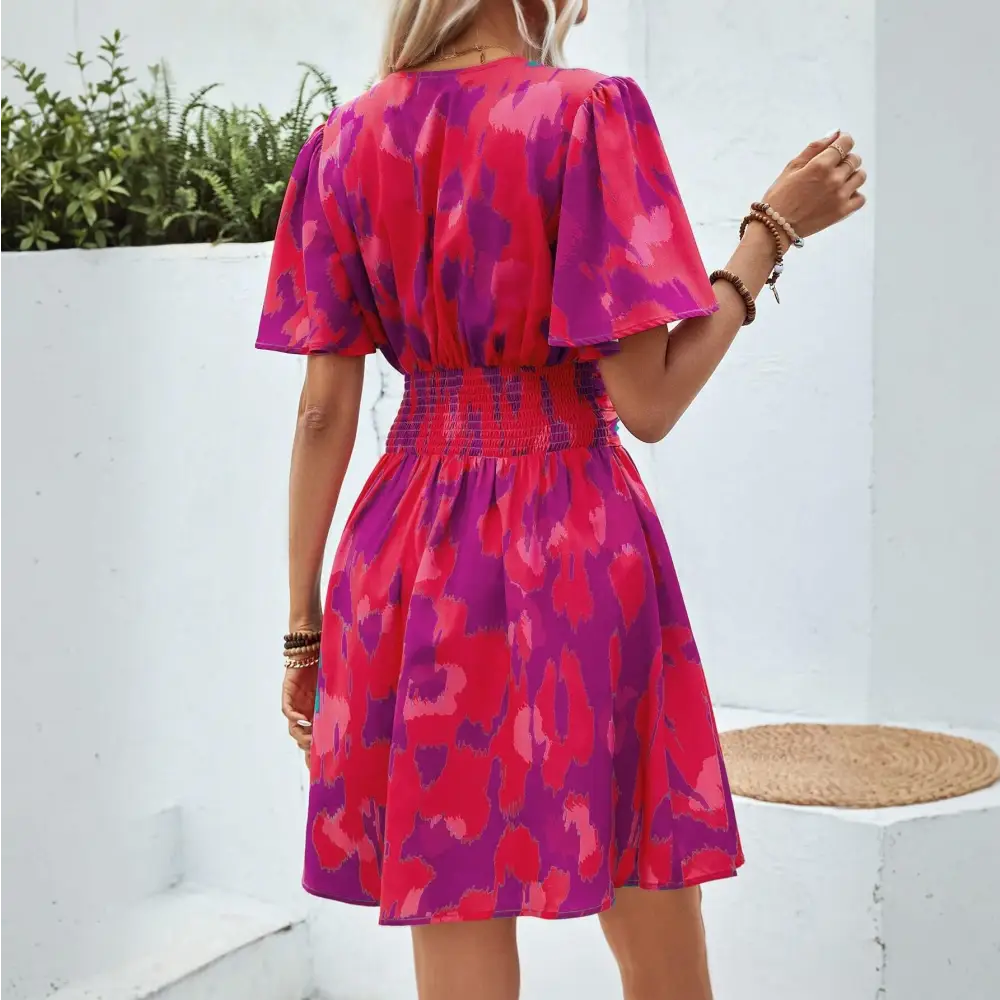 Vue de dos d'une robe cocktail chic et originale, avec un motif abstrait rose et violet, une ceinture élastique à la taille et des manches courtes flottantes, portée par une femme blonde.
