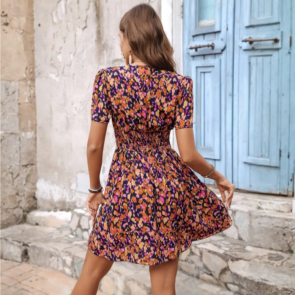 Vue de dos d'une robe cocktail courte à motifs floraux colorés, avec un dos smocké et des manches courtes, portée par une femme dans un cadre extérieur pittoresque.