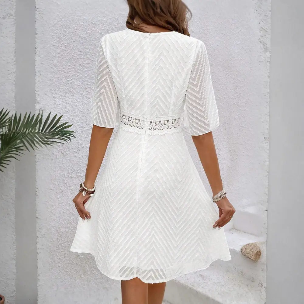 Une robe cocktail blanche en dentelle bohème chic, vue de dos, montrant le détail du tissu en dentelle avec des motifs géométriques. La ceinture en dentelle à la taille est visible.