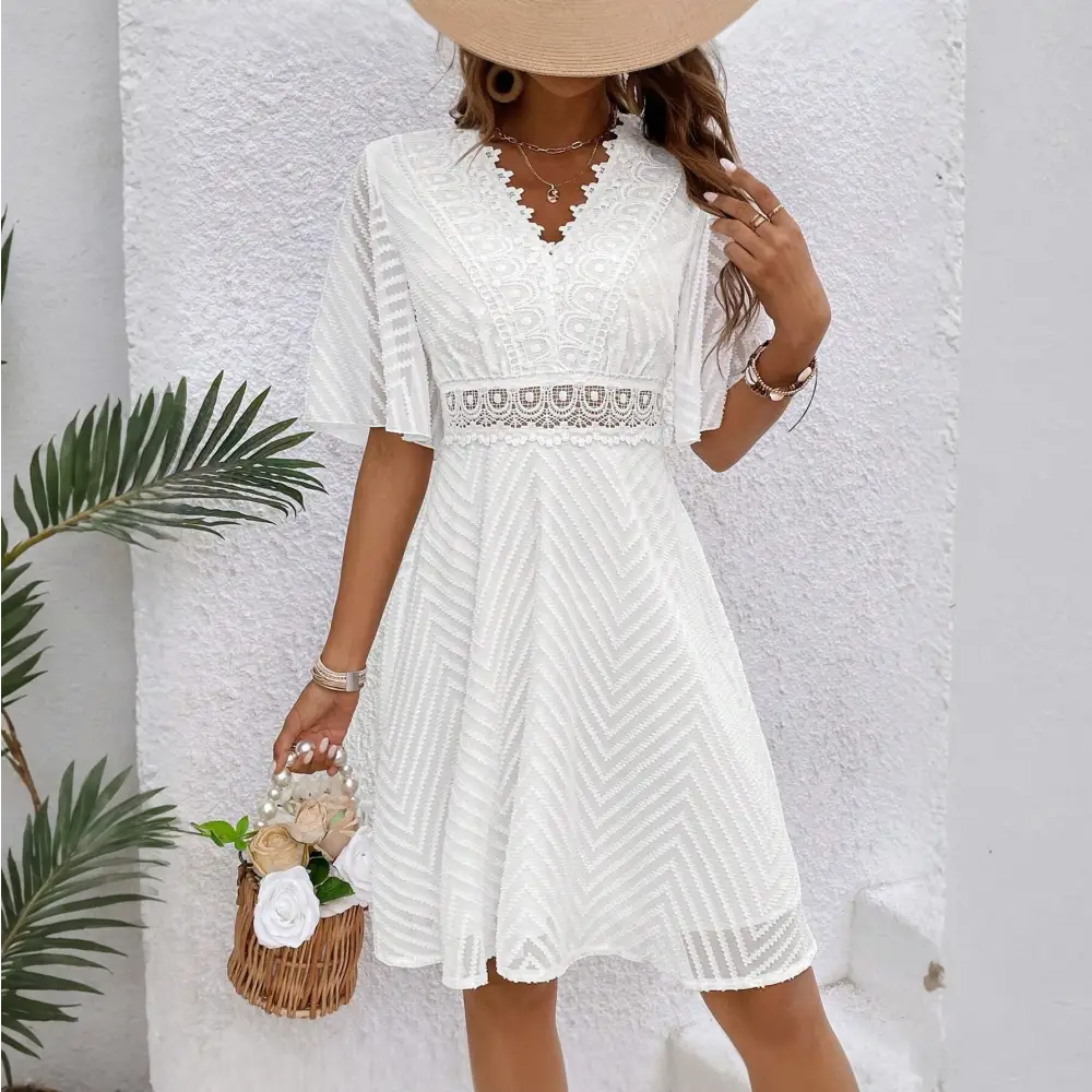 Une robe cocktail blanche en dentelle bohème chic, vue de face, portée avec un grand chapeau beige et un sac en osier rempli de fleurs blanches. Le décolleté en V et la ceinture en dentelle accentuent la taille.
