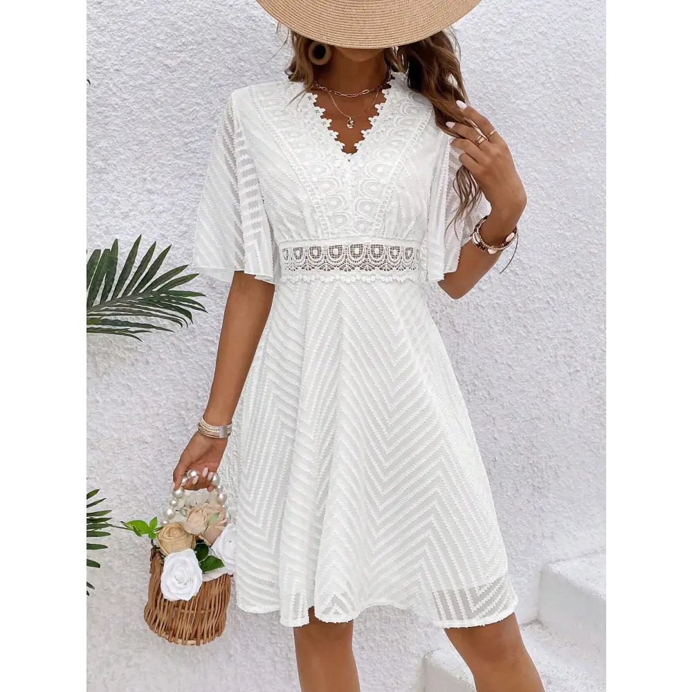 Une robe cocktail blanche en dentelle bohème chic, vue de face, portée avec un grand chapeau beige et un sac en osier rempli de fleurs blanches. Le décolleté en V et la ceinture en dentelle accentuent la taille.