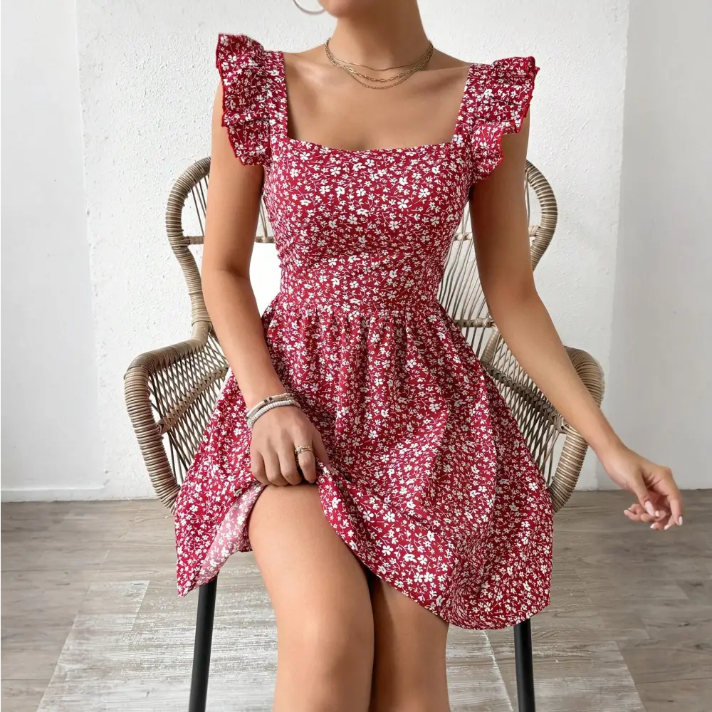 Une femme est assise sur une chaise en rotin, portant une robe cocktail rouge à motifs floraux blancs. La robe a des manches à volants et un décolleté carré. La femme tient légèrement le bas de la robe tout en étant assise.