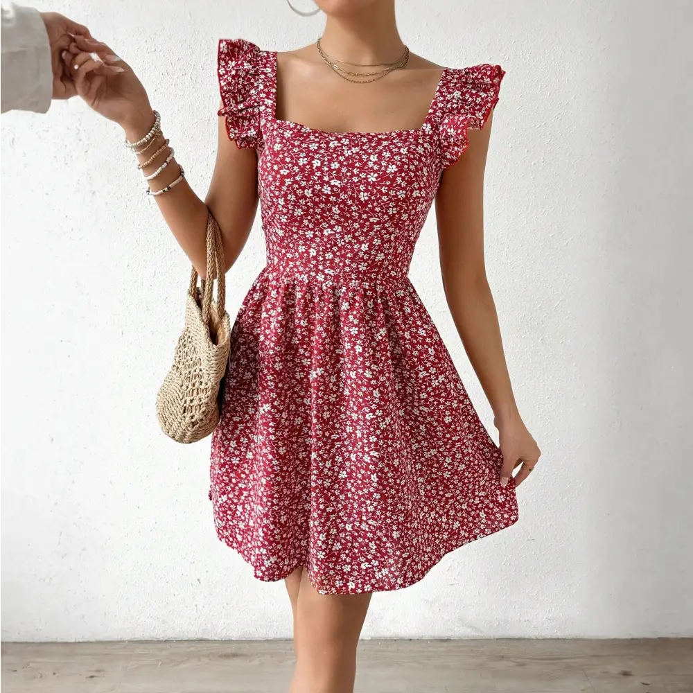 Une femme porte une robe cocktail rouge à motifs floraux blancs avec des manches à volants et un décolleté carré. Elle tient un sac en paille tressée à la main gauche et arbore plusieurs bracelets au poignet droit.
