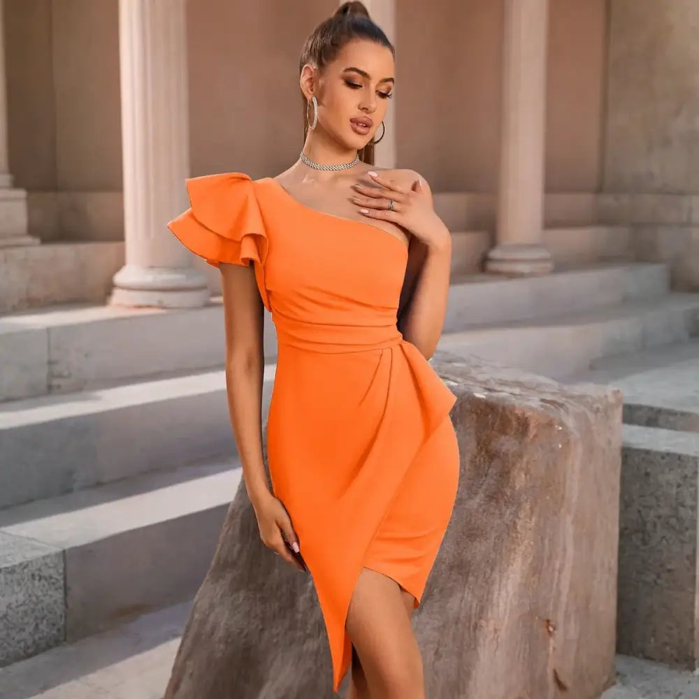 Une élégante robe cocktail orange portée par un mannequin. La robe est asymétrique avec une seule épaule ornée d’un grand volant. La coupe ajustée souligne la silhouette et présente une fente sur le devant. Le mannequin est adossé à un rocher, ajoutant une touche naturelle et contrastée à la scène. Elle porte des sandales transparentes et des bijoux dorés, complétant un look chic et estival.