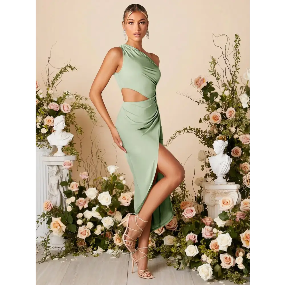 Femme portant une robe cocktail vert sauge, élégante et moderne, avec une épaule découverte et une découpe à la taille. La robe présente une fente haute à l'avant, ajoutant une touche de sensualité, et est ajustée pour mettre en valeur la silhouette. La femme est accessoirisée avec des boucles d'oreilles scintillantes et porte des sandales à talons. Elle est entourée de décorations florales, créant une ambiance parfaite pour un événement formel ou une soirée chic.