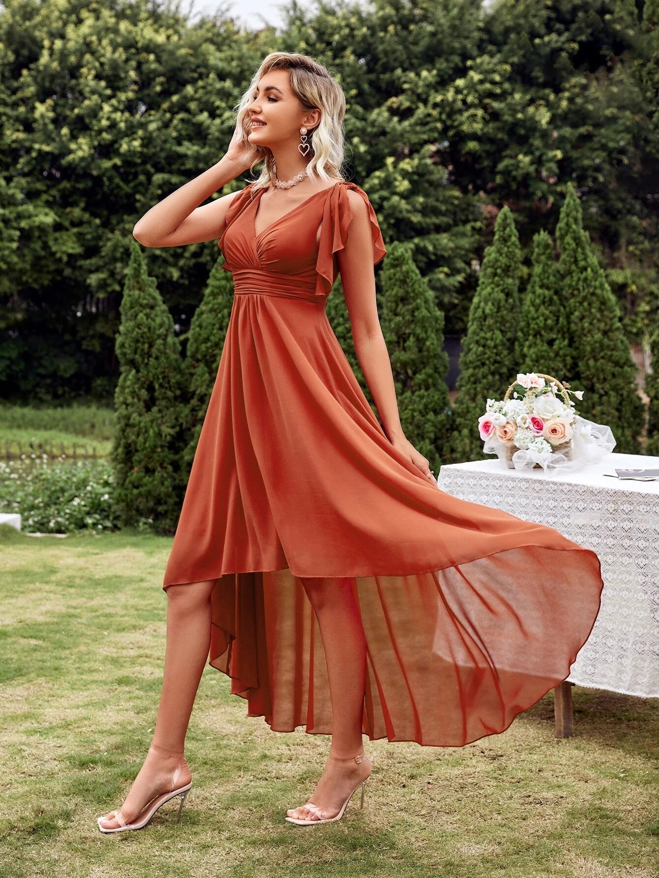 Vue de côté d'une robe cocktail chic originale en terracotta, avec un décolleté en V et des manches courtes, portée par une femme blonde dans un jardin, accompagnée de talons transparents.