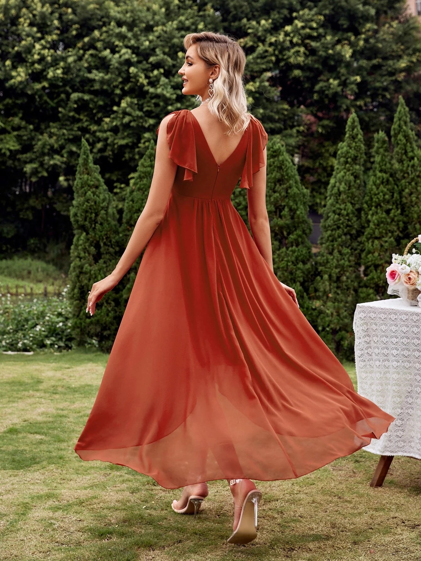 Vue de dos d'une élégante robe cocktail chic originale en terracotta, avec un décolleté en V et une coupe fluide, portée par une femme blonde dans un jardin verdoyant.