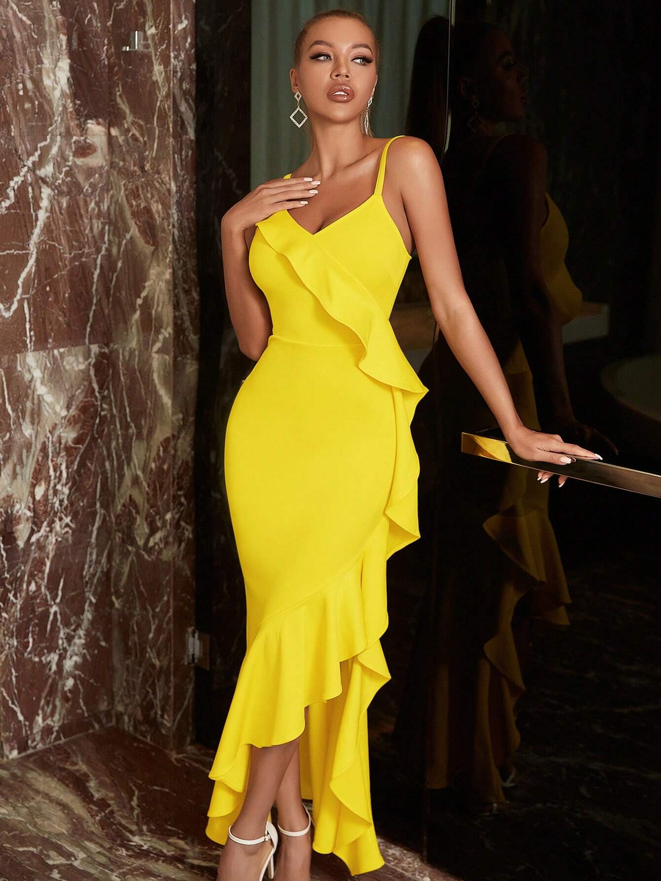 robe cocktail jaune elegante porte par un mannequin. La robe est sans manches avec des bretelles fines et un décolleté en V. La coupe moulante épouse la silhouette et est ornée de volants asymétriques qui ajoutent du mouvement. Le mannequin porte des boucles d’oreilles pendantes et des sandales à talons blancs, parfaites pour une soirée estivale.