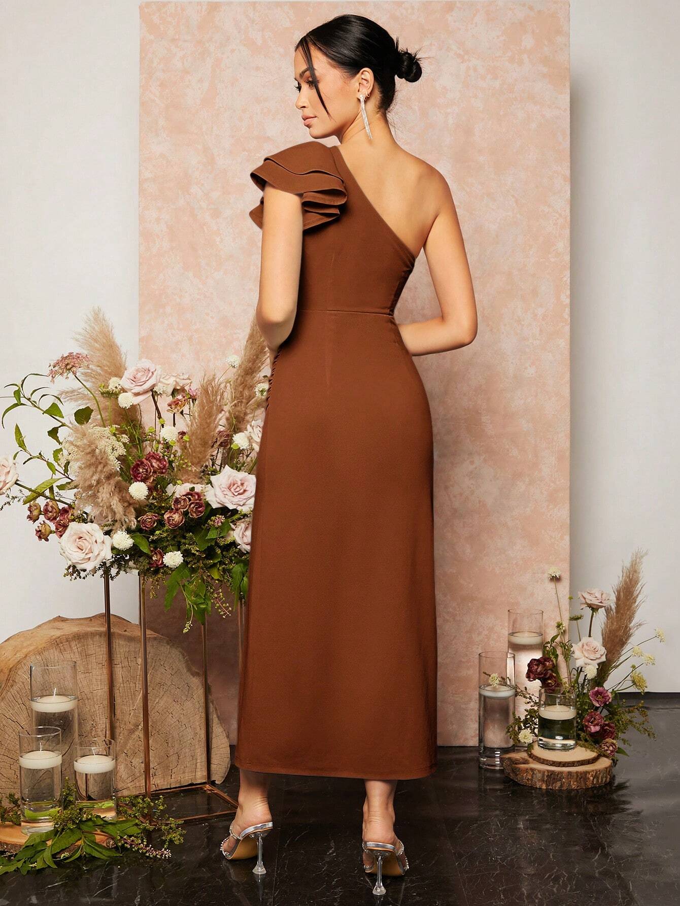 Vue arrière d’une élégante robe cocktail terracotta portée par un mannequin. La robe est asymétrique avec une seule épaule ornée d’un détail volanté. La coupe ajustée met en valeur la silhouette, tandis que le dos est découvert, ajoutant une touche de sophistication. Le décor floral avec des fleurs et des bougies crée une ambiance romantique.