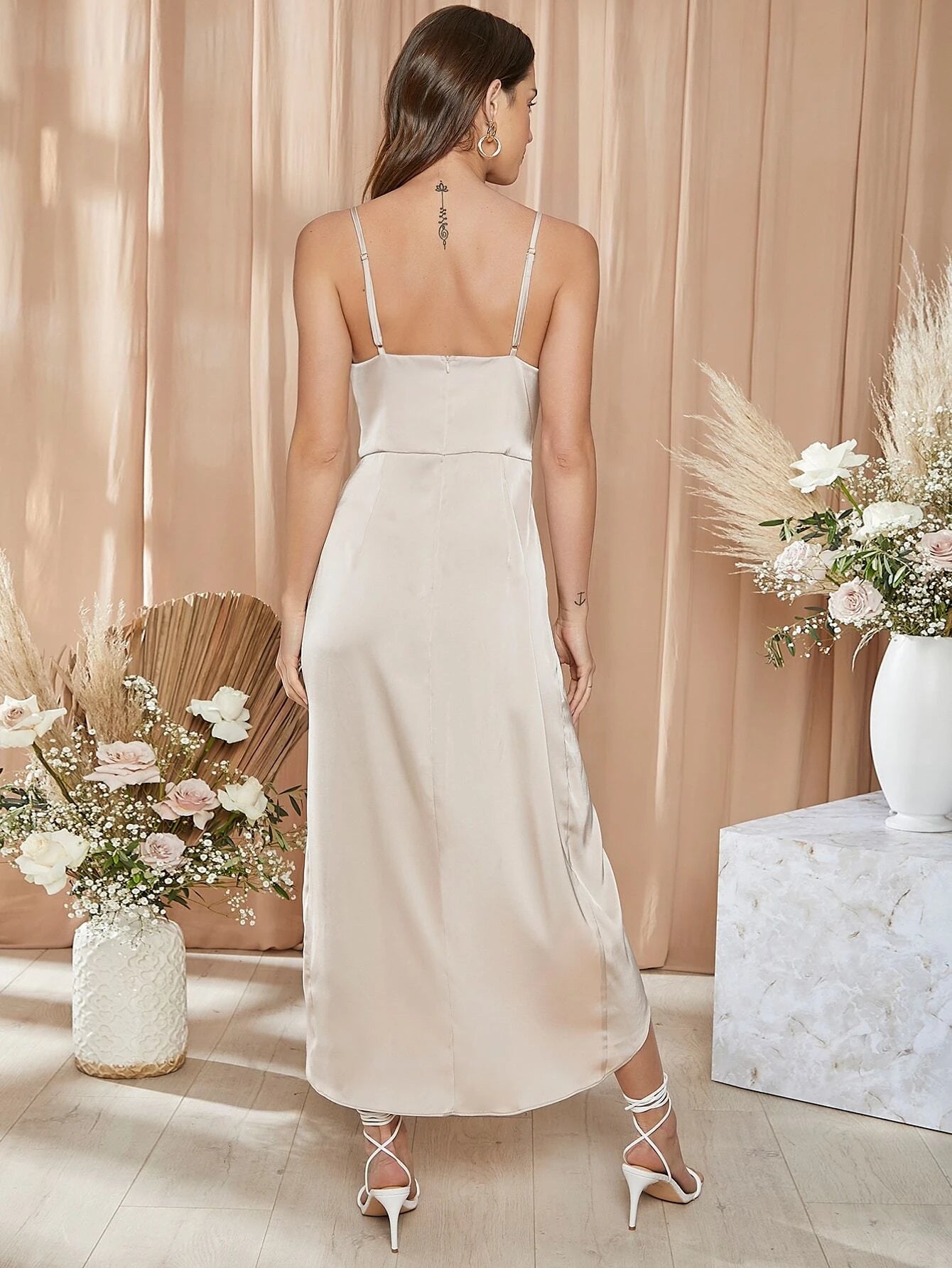 Vue de dos d'une robe de mariée cocktail en satin beige, avec des bretelles fines et une coupe élégante, portée par une femme aux cheveux bruns dans un décor floral sophistiqué.