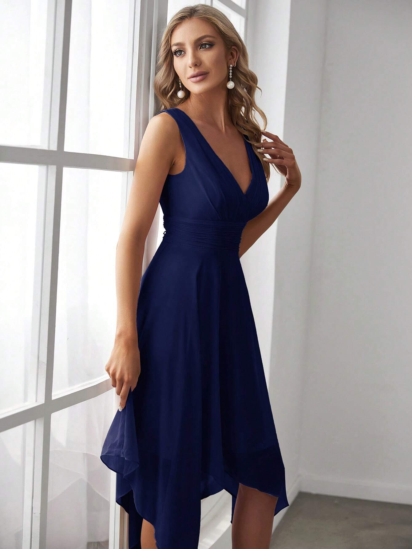 Femme portant une robe de cocktail bleue pour mariage, adossée à une fenêtre, vue de profil, montrant les détails plissés de la taille.