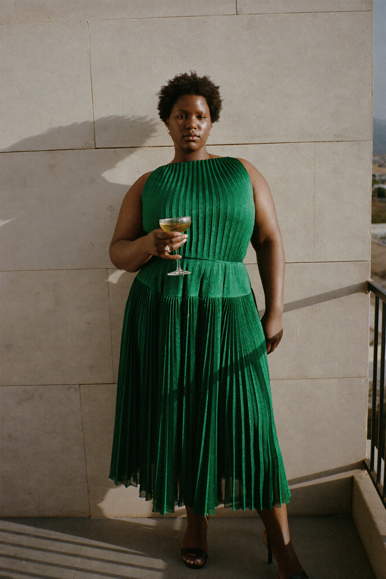 Robe cocktail émeraude plissée, coupe de champagne à la main, terrasse au soleil couchant