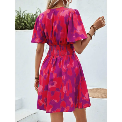 Vue de dos d'une robe cocktail chic et originale, avec un motif abstrait rose et violet, une ceinture élastique à la taille et des manches courtes flottantes, portée par une femme blonde.
