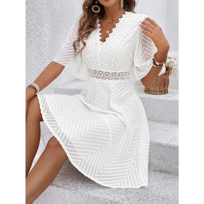 Une robe cocktail blanche en dentelle bohème chic, vue de face, assise sur des escaliers. Le décolleté en V et la ceinture en dentelle sont bien mis en valeur, créant un look chic et estival.