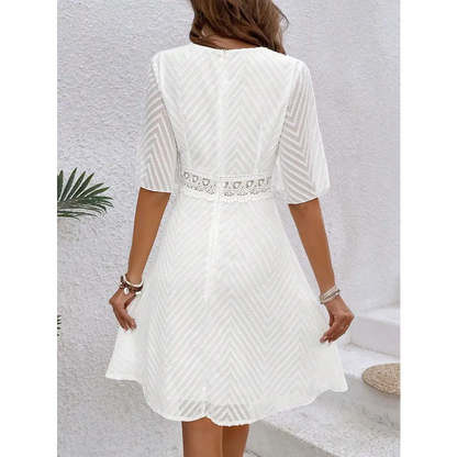 Une robe cocktail blanche en dentelle bohème chic, vue de dos, montrant le détail du tissu en dentelle avec des motifs géométriques. La ceinture en dentelle à la taille est visible.