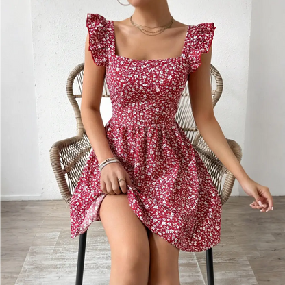 Une femme est assise sur une chaise en rotin, portant une robe cocktail rouge à motifs floraux blancs. La robe a des manches à volants et un décolleté carré. La femme tient légèrement le bas de la robe tout en étant assise.