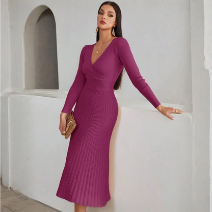 Image d’une femme posant de manière élégante dans une robe de cocktail hiver en tricot violet. La robe présente un décolleté en V, des manches longues et une jupe plissée. La femme est accessoirisée avec des boucles d’oreilles dorées et un sac à main beige, et elle porte des bottes blanches. Elle pose avec une main sur la hanche, dans un intérieur minimaliste.
