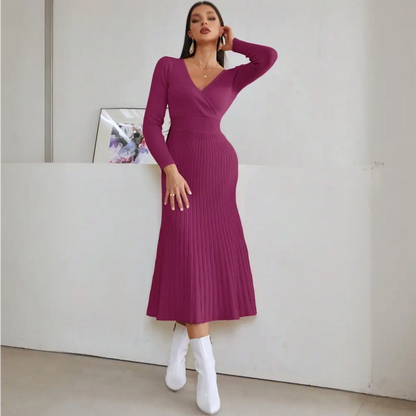 	Image d’une femme portant une élégante robe de cocktail hiver en tricot violet. La robe est à manches longues, avec un décolleté en V et une jupe plissée qui tombe jusqu’à mi-mollet. La femme porte des boucles d’oreilles dorées et tient un sac à main beige. Elle pose de face, appuyée contre un mur blanc.