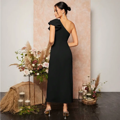 Vue arrière de la robe cocktail noire. La robe montre un dos découvert avec une épaule nue, accentuant la féminité et l’élégance. Le détail volanté sur l’épaule unique ajoute du charme, tandis que la coupe ajustée met en valeur la silhouette. Le décor avec des fleurs et des bougies crée une ambiance romantique et sophistiquée.