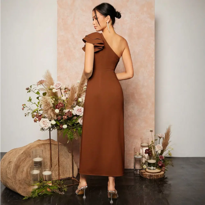 Vue arrière d’une élégante robe cocktail terracotta portée par un mannequin. La robe est asymétrique avec une seule épaule ornée d’un détail volanté. La coupe ajustée met en valeur la silhouette, tandis que le dos est découvert, ajoutant une touche de sophistication. Le décor floral avec des fleurs et des bougies crée une ambiance romantique.