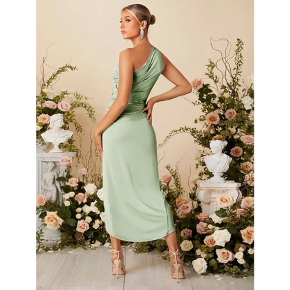 Vue arrière d'une femme portant une robe cocktail vert sauge, élégante et moderne, avec une épaule découverte. La robe est ajustée pour mettre en valeur la silhouette et présente une longueur mi-mollet. La femme est accessoirisée avec des boucles d'oreilles scintillantes et porte des sandales à talons. Elle est entourée de décorations florales, créant une ambiance parfaite pour un événement formel ou une soirée chic.