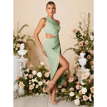 Femme portant une robe cocktail vert sauge, élégante et moderne, avec une épaule découverte et une découpe à la taille. La robe présente une fente haute à l'avant, ajoutant une touche de sensualité, et est ajustée pour mettre en valeur la silhouette. La femme est accessoirisée avec des boucles d'oreilles scintillantes et porte des sandales à talons. Elle est entourée de décorations florales, créant une ambiance parfaite pour un événement formel ou une soirée chic.
