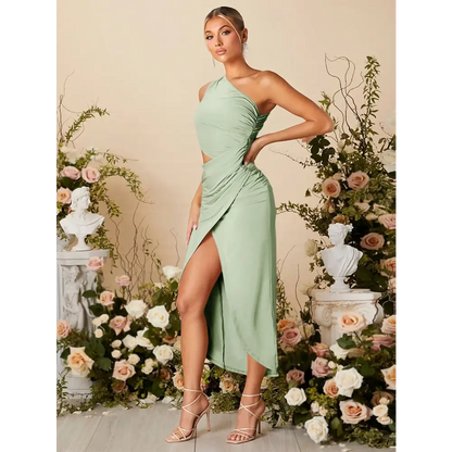 Vue latérale d'une femme portant une robe cocktail vert sauge, élégante et moderne, avec une épaule découverte et une découpe à la taille. La robe présente une fente haute à l'avant, ajoutant une touche de sensualité, et est ajustée pour mettre en valeur la silhouette. La femme est accessoirisée avec des boucles d'oreilles scintillantes et porte des sandales à talons. Elle est entourée de décorations florales, créant une ambiance parfaite pour un événement formel ou une soirée chic.